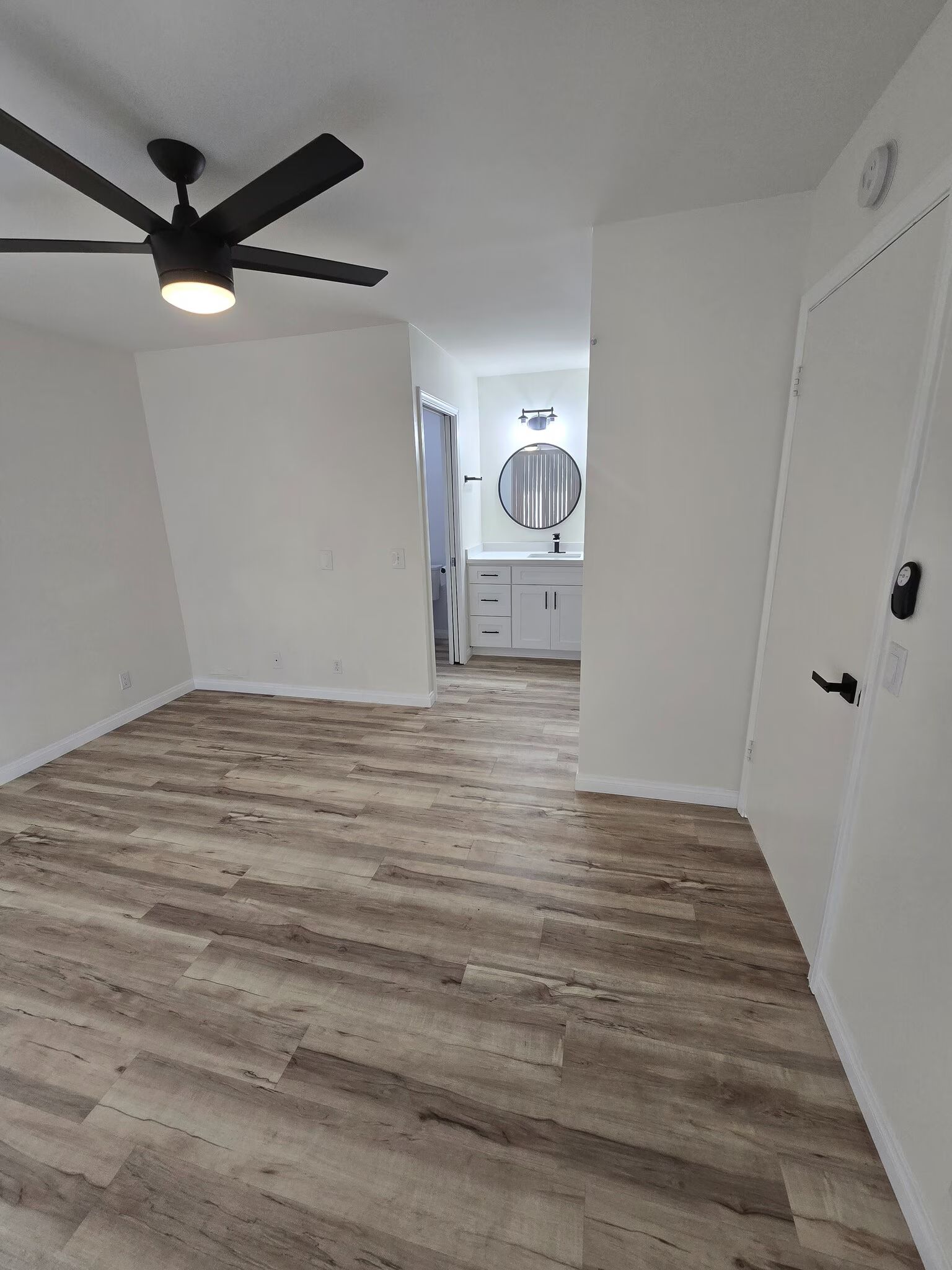 Empty modern room with wood-look flooring, white walls, black ceiling fan, and a doorway to a bright bathroom.