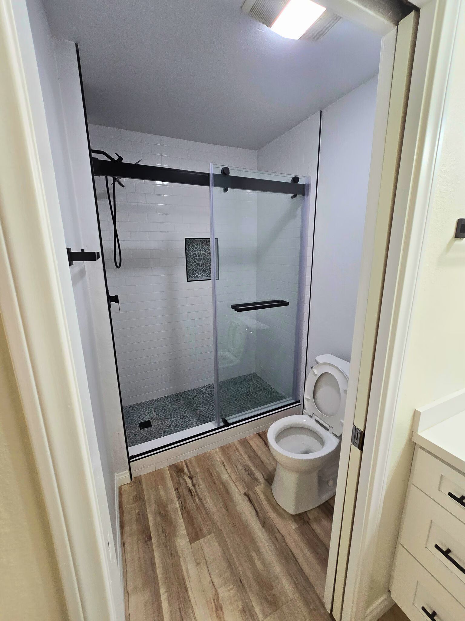 Small bathroom with glass shower, wood-look floor, and toilet beside a vanity cabinet.