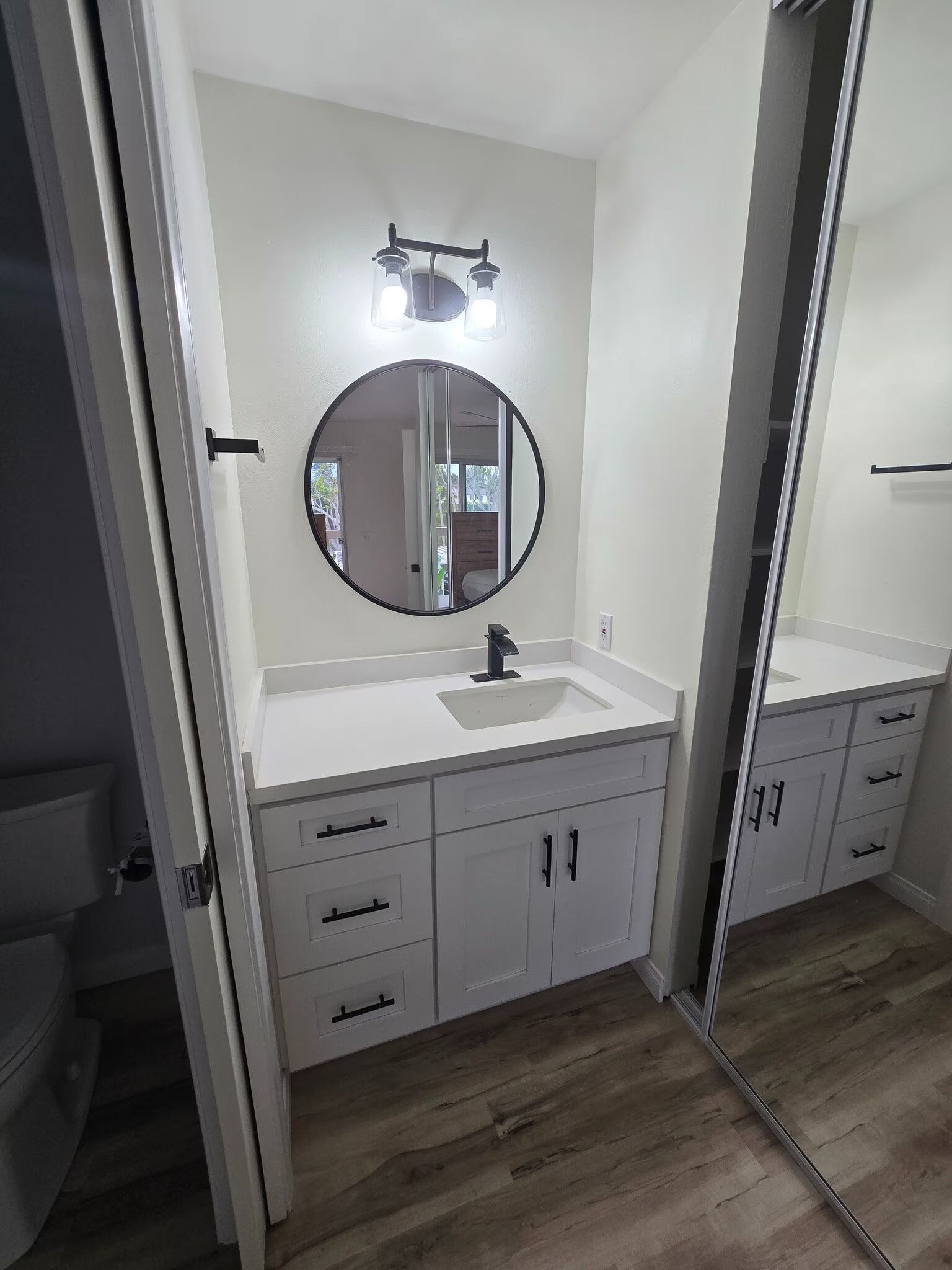 Small bathroom vanity with round mirror, black faucet, white cabinets, and wood-look floor.