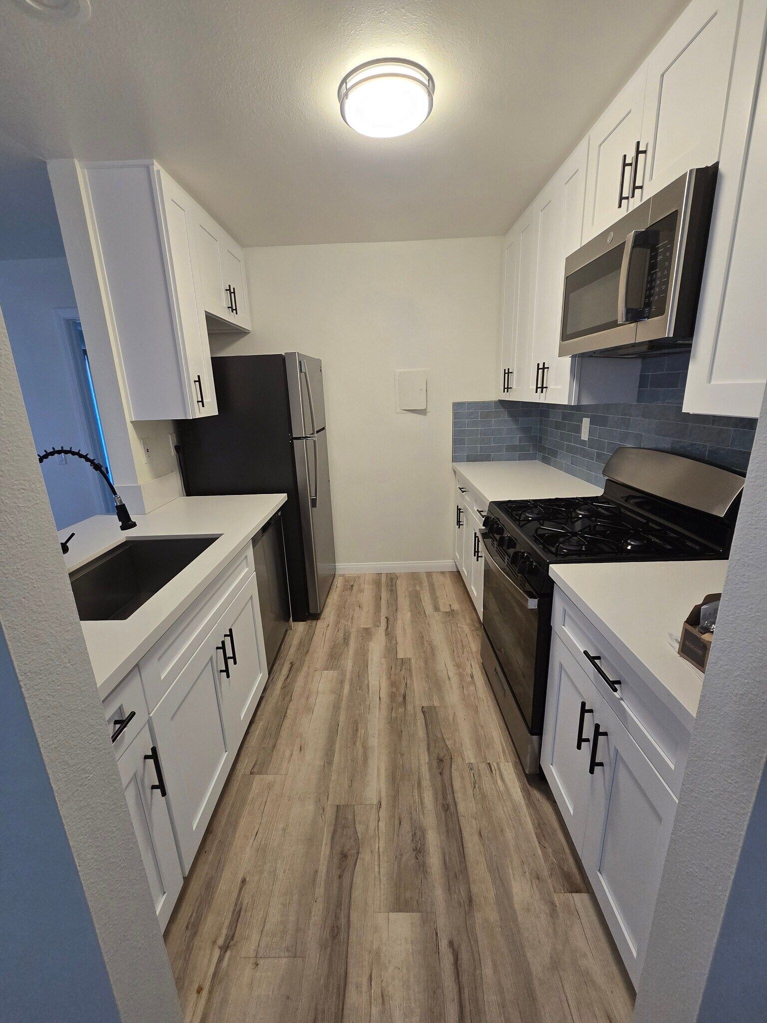 Narrow modern kitchen with white cabinets, black countertops, stainless appliances, and wood-look flooring.