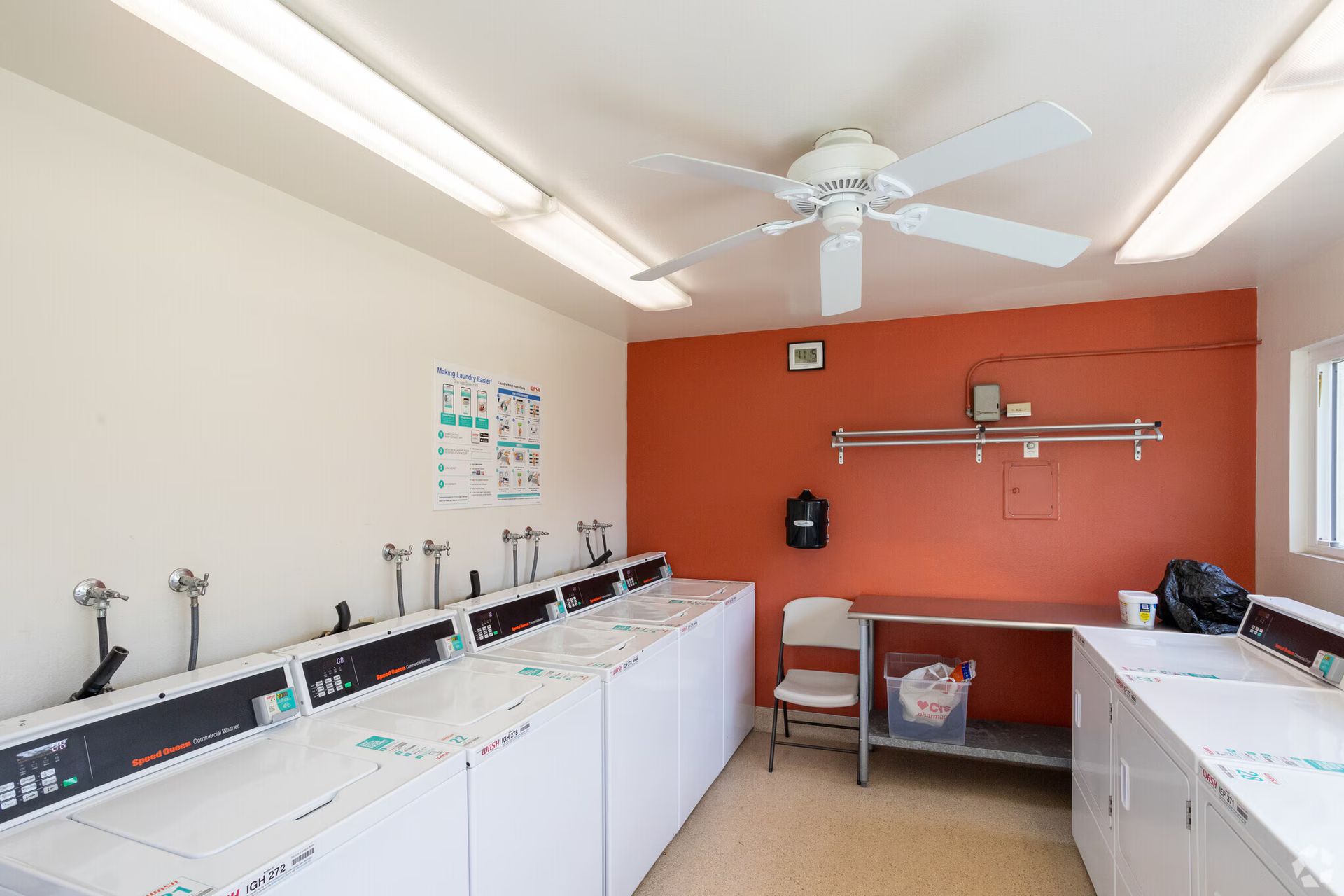 Laundry room with rows of washers and dryers, white walls, and a red accent wall with a ceiling fan.