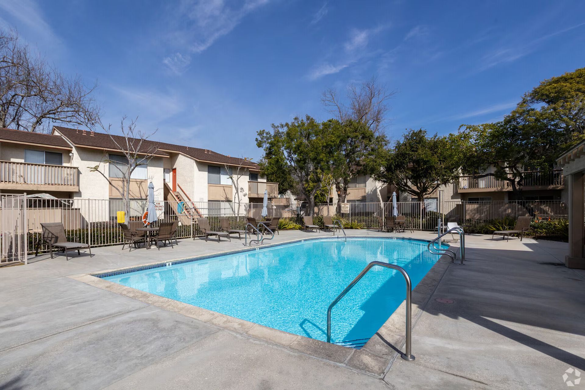 Apartment complex pool with blue water, lounge chairs, and trees under a clear sky