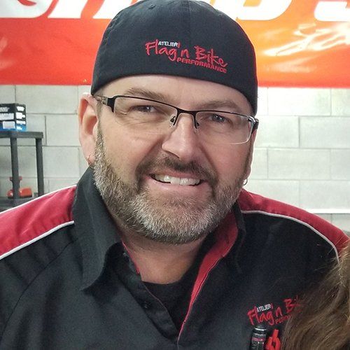 Homme avec des lunettes et une barbe portant une casquette noire et une chemise de travail, souriant à la caméra.