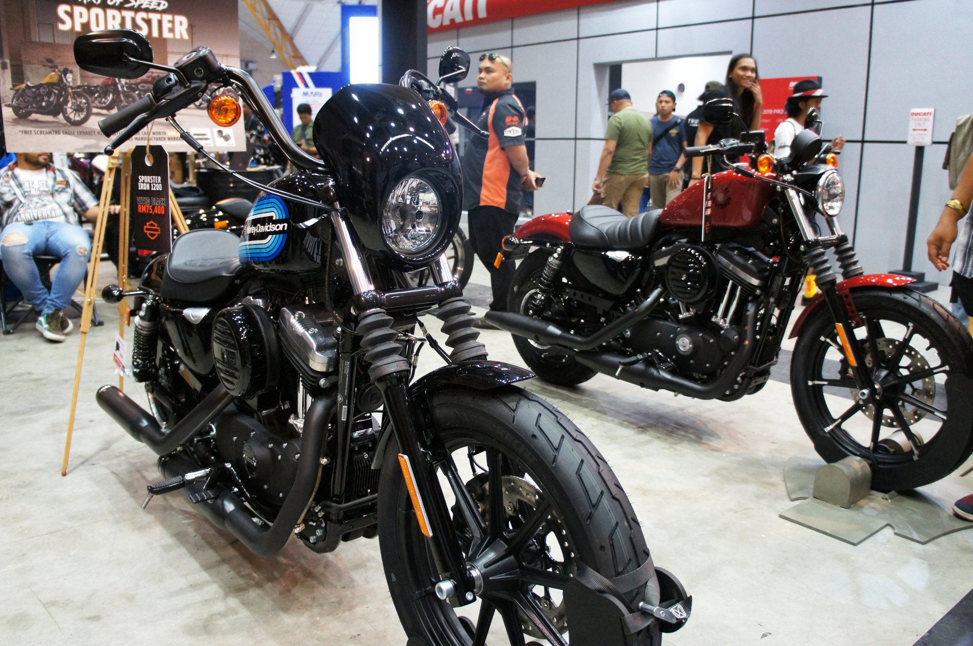 Deux Harley-Davidson exposées lors d'un événement en intérieur ; l'une noire, l'autre rouge. Des personnes sont visibles à l'arrière-plan.