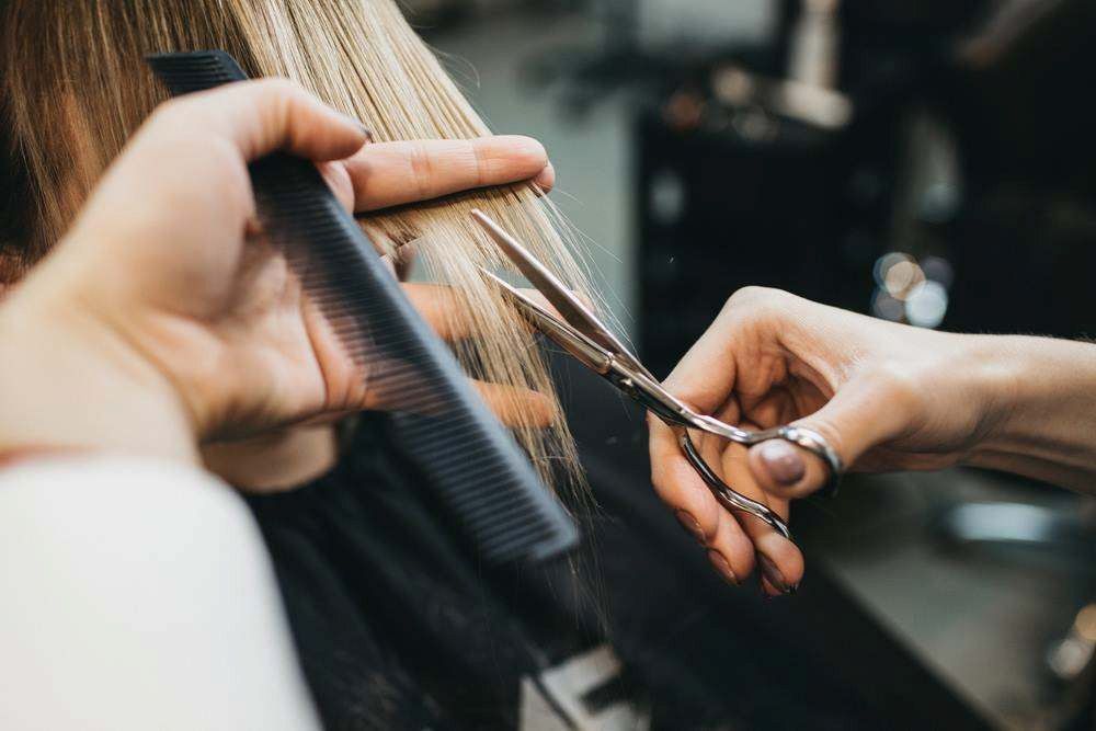 Hands Cutting Long Blonde Hair With Scissors and a Comb in a Salon — Elysian Salon In Wyong, NSW