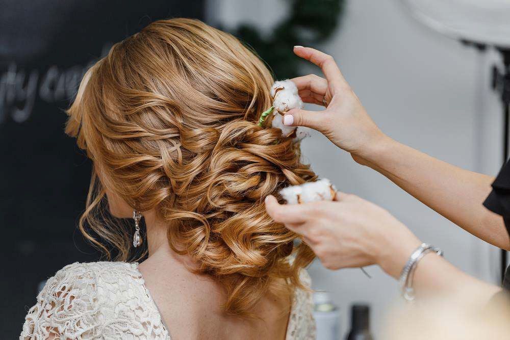 Stylist Arranging an Elegant Braided Updo With Floral Hairpin on a Woman in a Lace Dress — Elysian Salon In Wyong, NSW