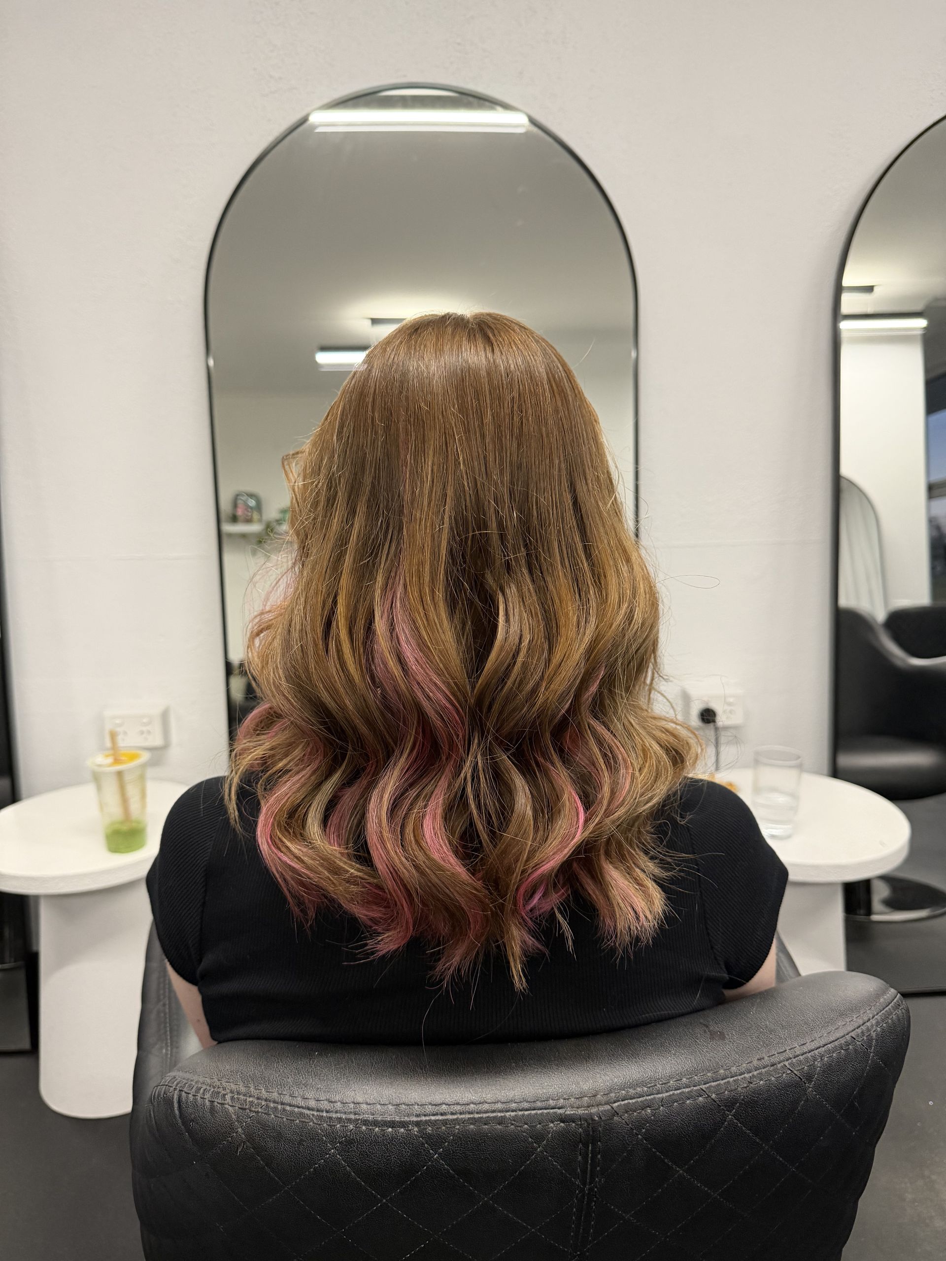 A Woman is Sitting in a Chair in Front of a Sign That Says Cherry Bears — Choo Choo Hair and Beauty In Wyong, NSW Person with wavy brown-and-pink hair seated in a salon chair, seen from behind between mirrors — Elysian Salon In Wyong, NSW