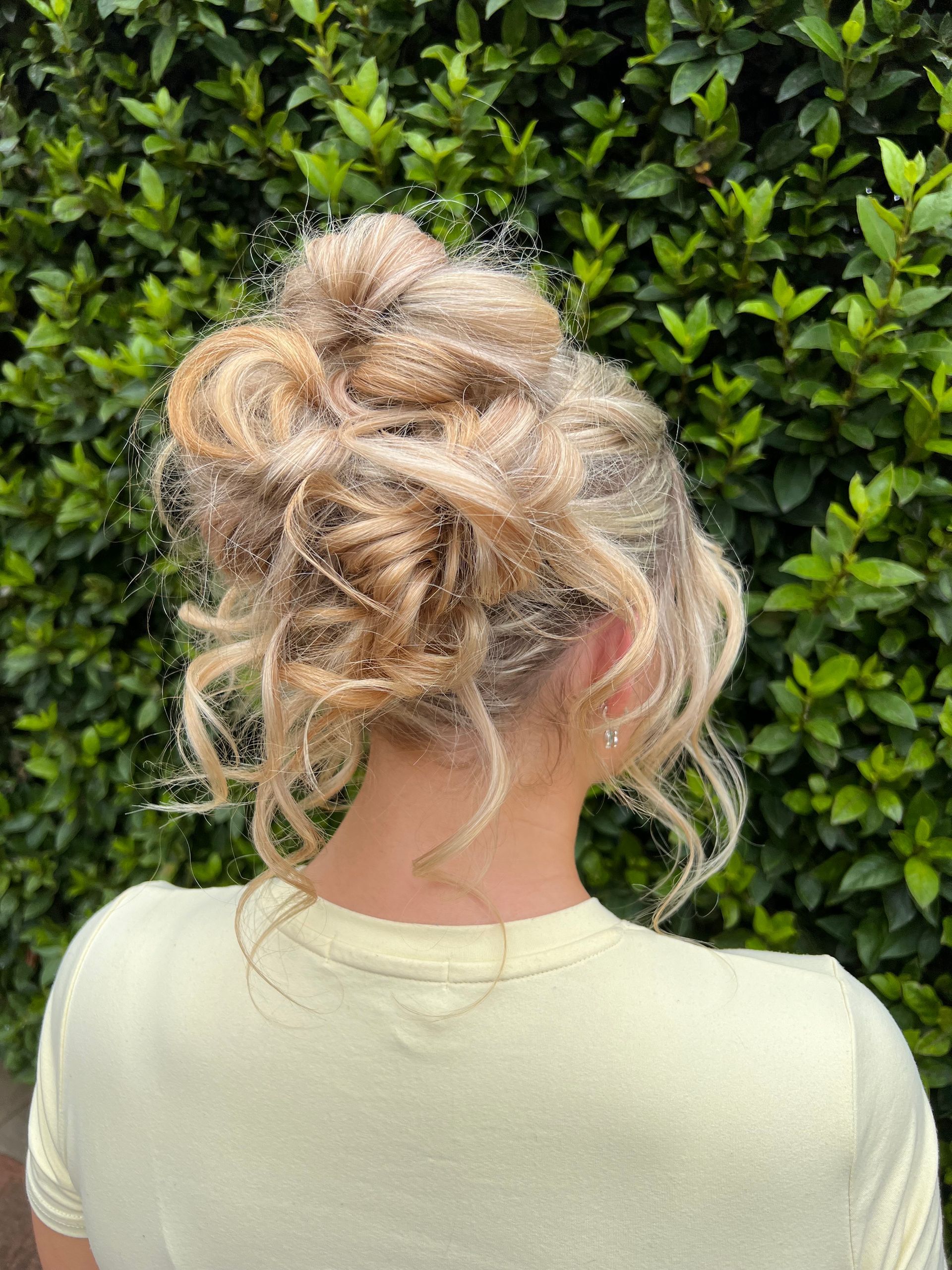 Blonde curly hair in a messy bun against a green leafy hedge, viewed from behind — Elysian Salon In Wyong, NSW