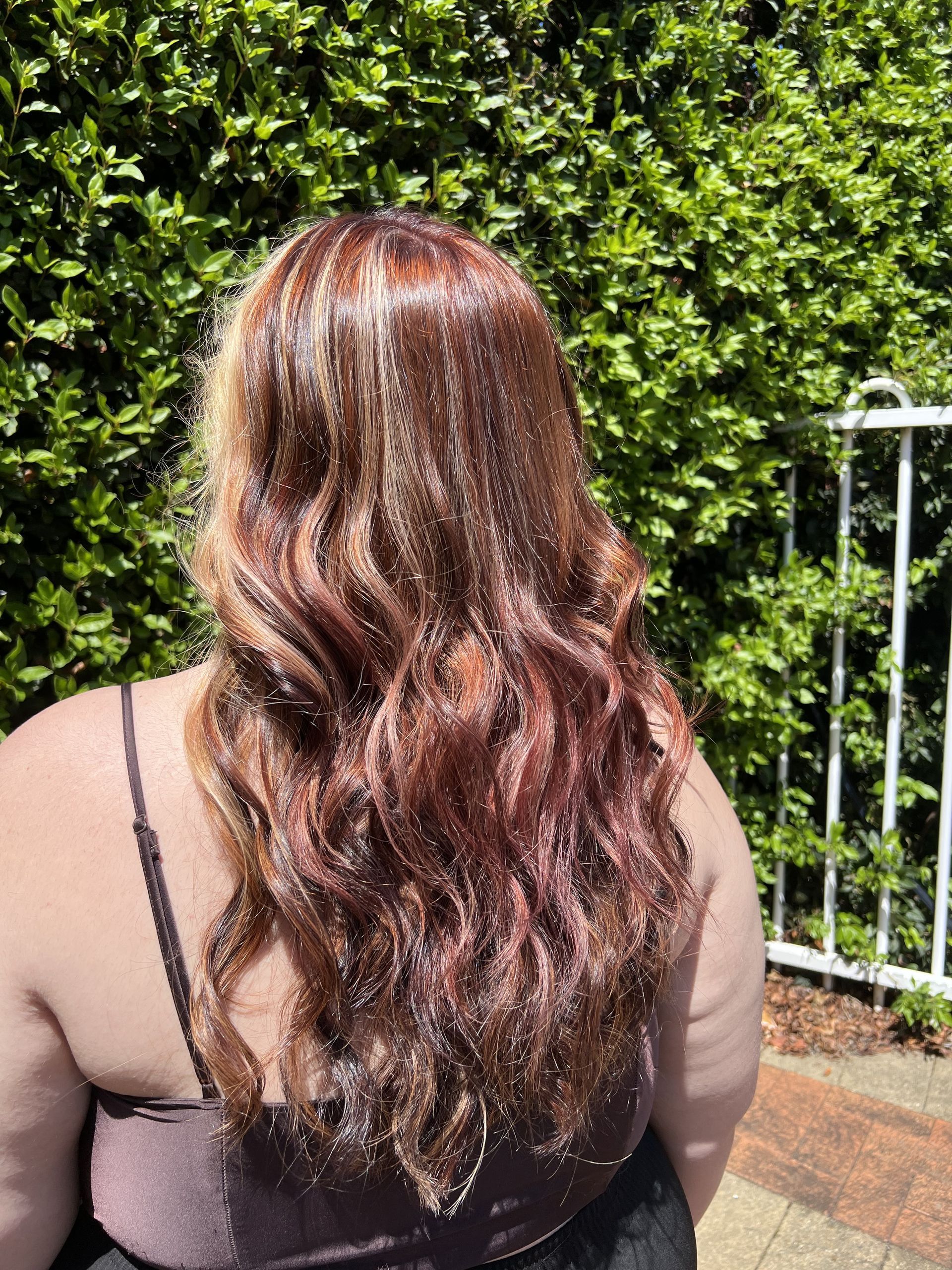 Person with long wavy brown hair seen from behind outdoors beside a green hedge and fence — Elysian Salon In Wyong, NSW