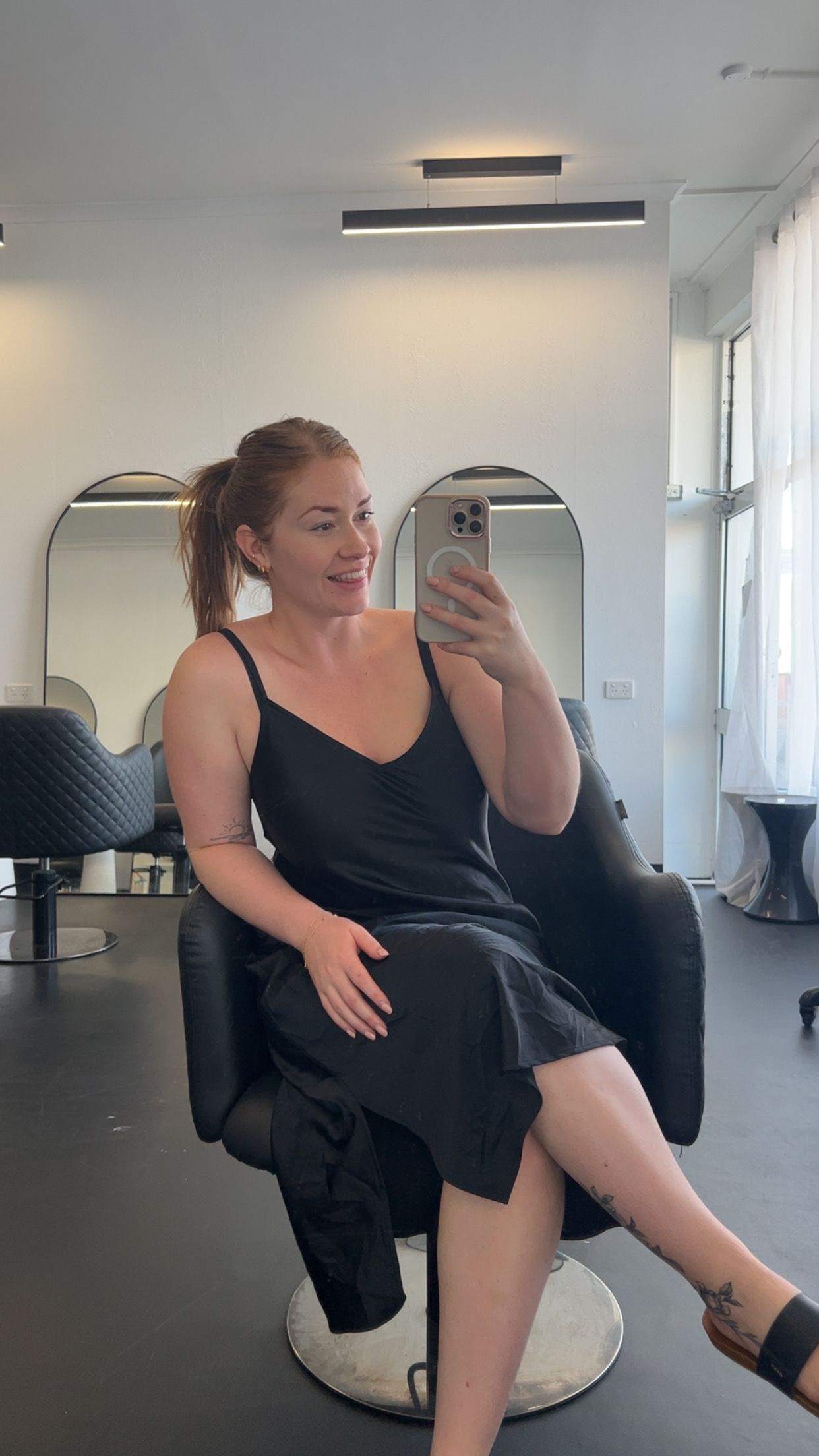 Woman in a black dress taking a mirror selfie in a bright room with a standing lamp and chairs — Elysian Salon In Wyong, NSW