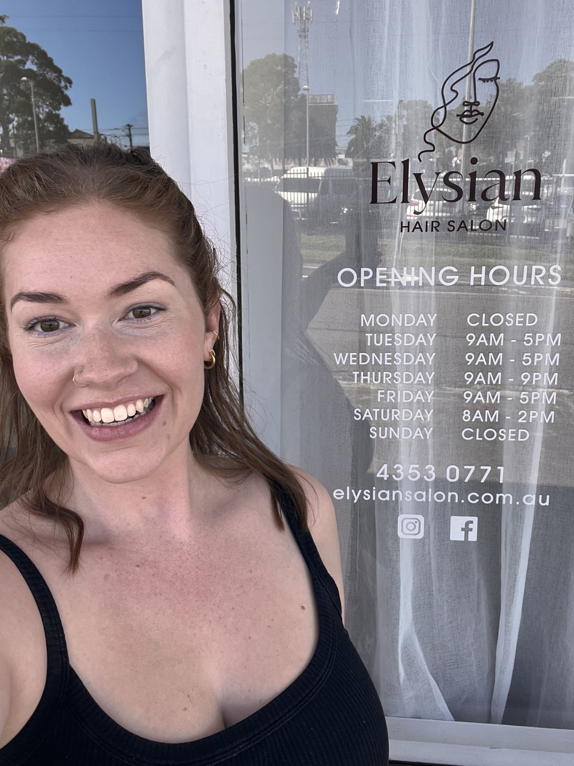 Smiling woman standing beside a salon window with business hours and logo signage visible — Elysian Salon In Wyong, NSW
