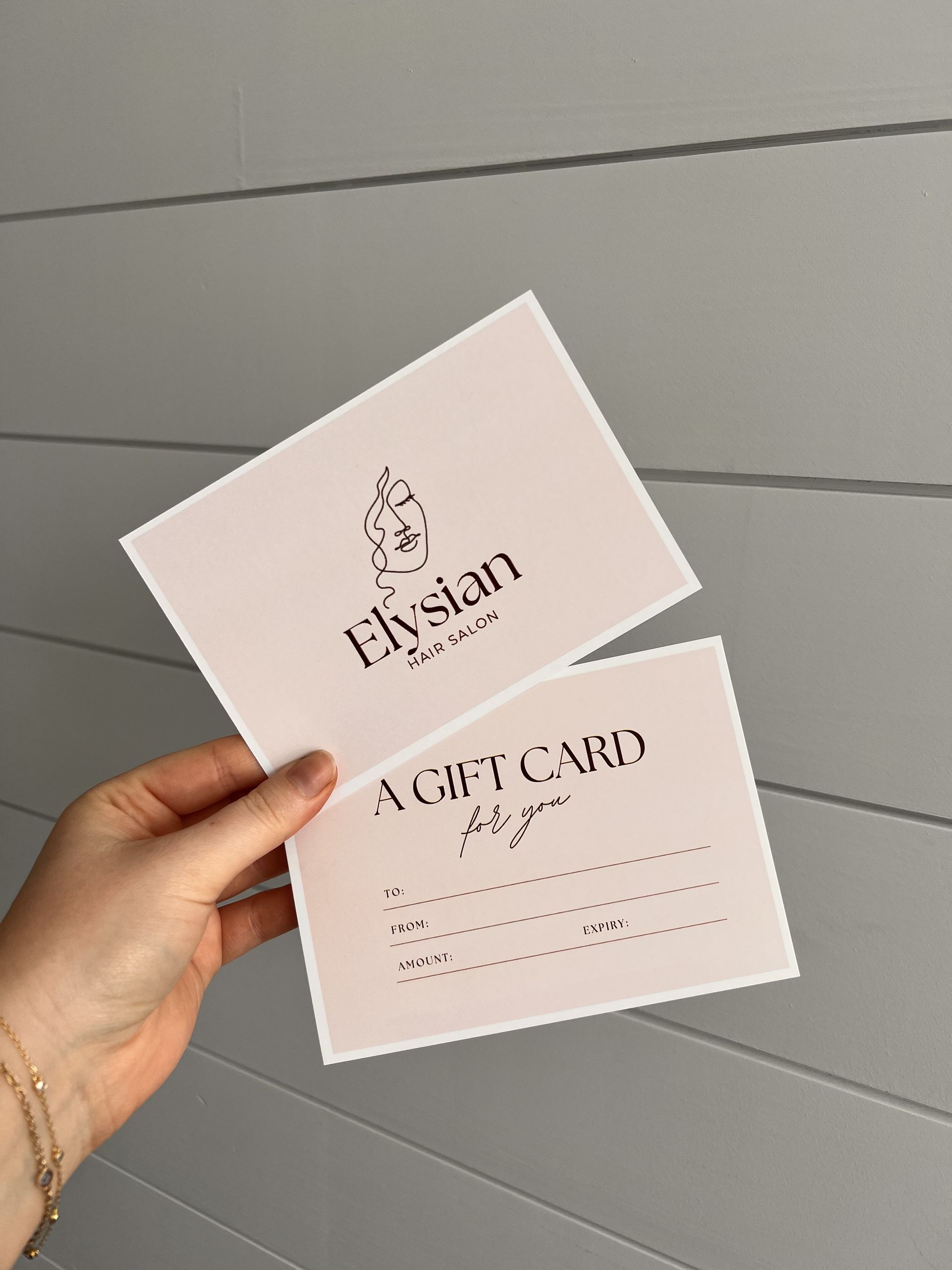 Hand holding two pink gift cards with the word “Elysian” against a gray wall — Elysian Salon In Wyong, NSW
