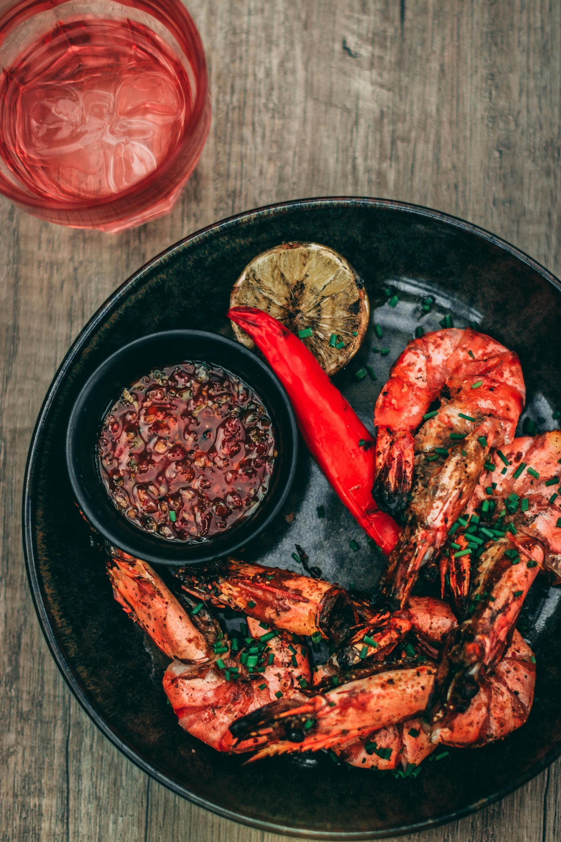 Grilled shrimp with dipping sauce, lime, chili pepper, and drink on a wood table.