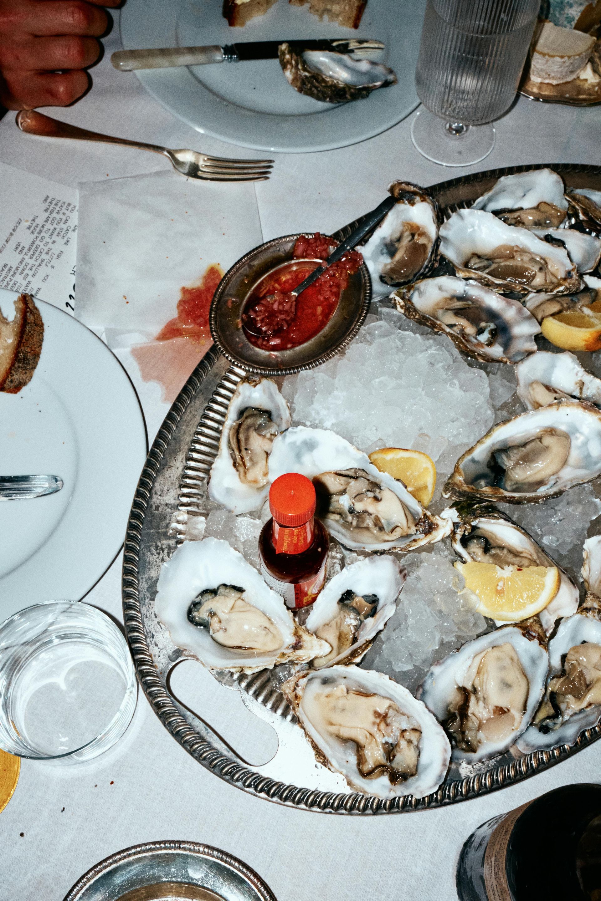 Oysters on ice on a silver platter with hot sauce and cocktail sauce, served at a table.