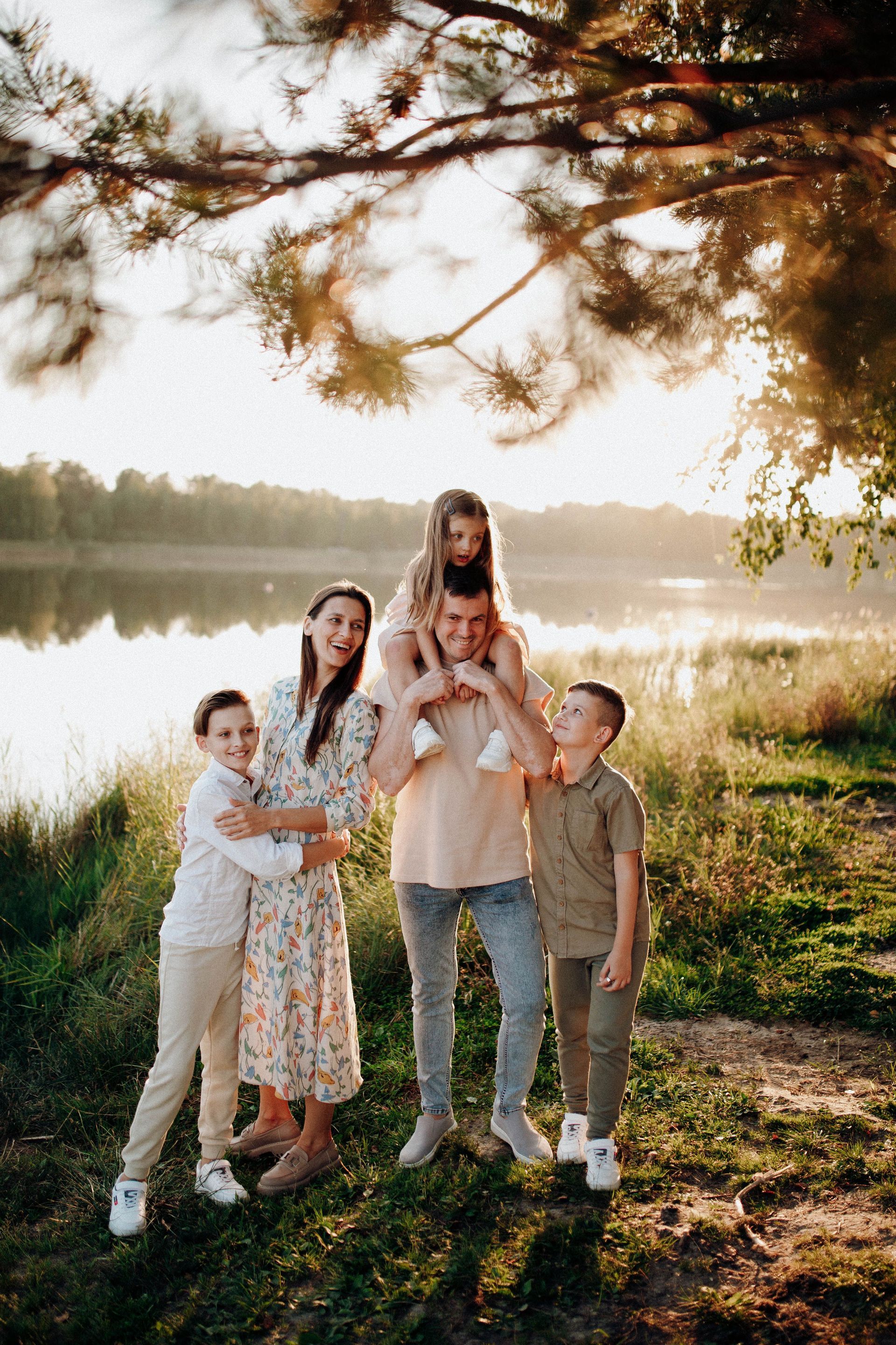 Una familia posando a la orilla de un lago al atardecer, con árboles sobre sus cabezas y una cálida luz dorada.