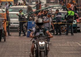 Un motociclista se dirige hacia la cámara en una calle concurrida, con agentes de policía y transeúntes detrás de barreras.