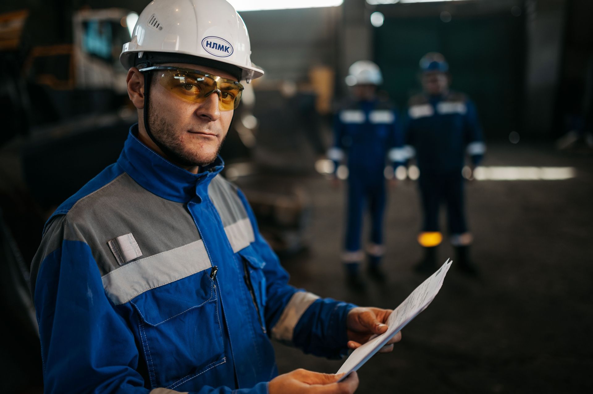 Trabajador con traje de seguridad azul y casco sosteniendo un documento en una instalación industrial.