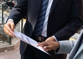 Dos personas vestidas con traje revisan un documento al aire libre, cerca de una barandilla.