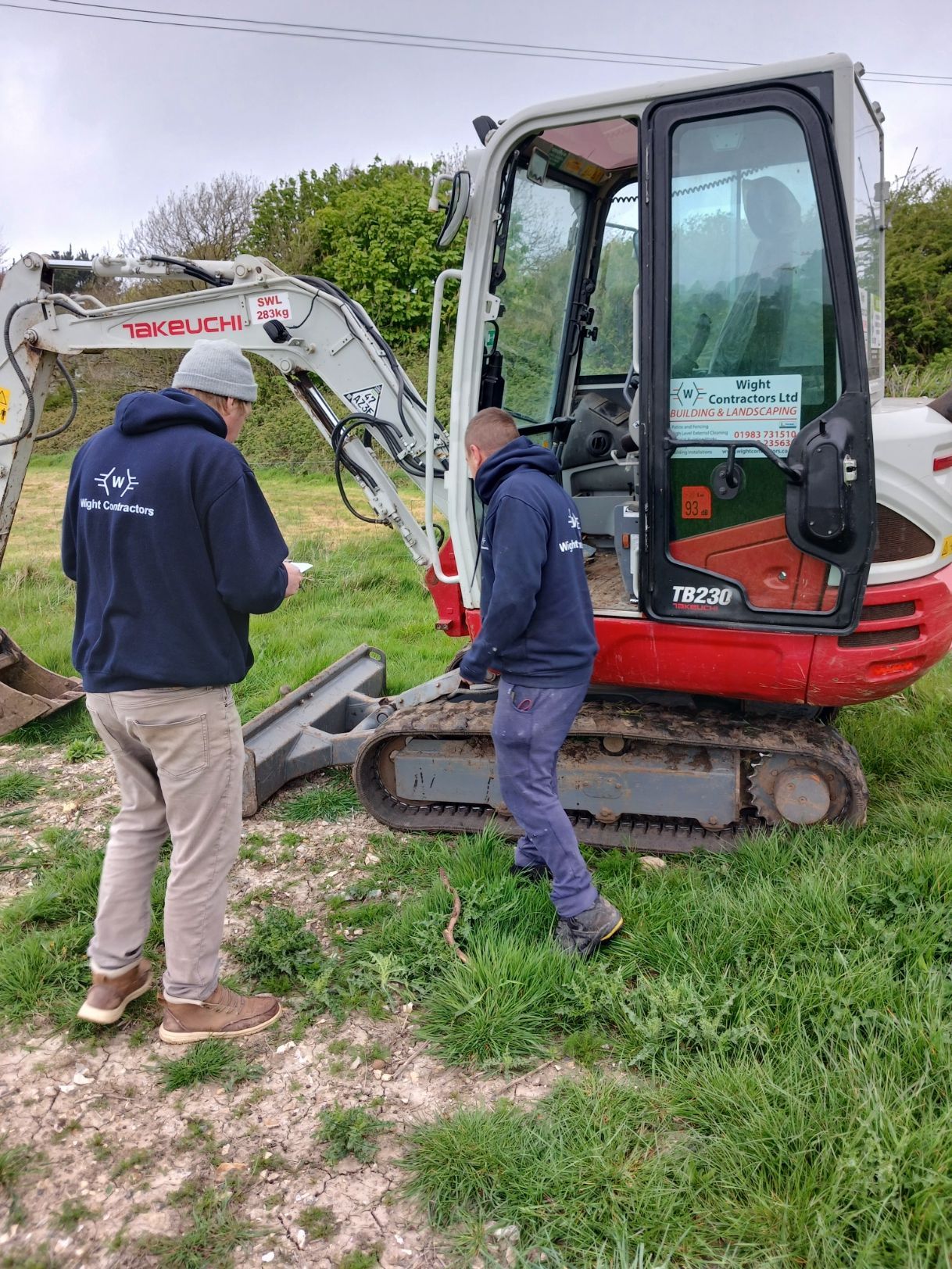 Two workers inspect a white mini excavator with a red cab in a grassy yard.