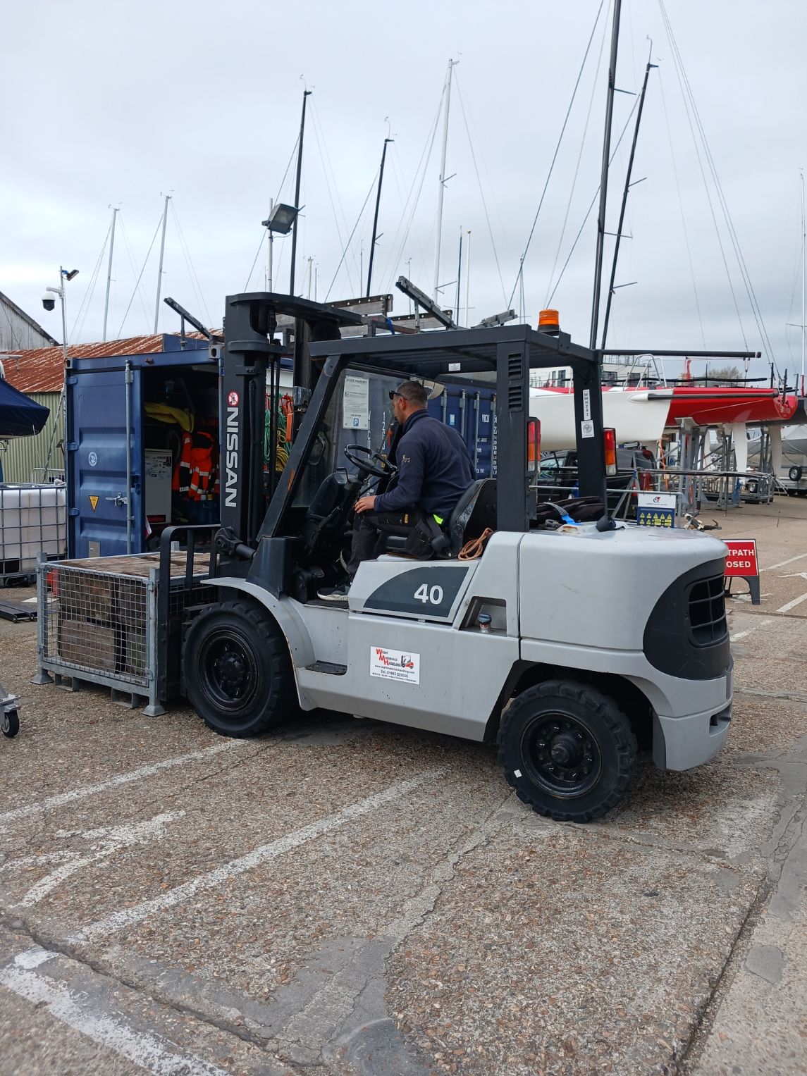 Forklift parked at a harbor dock, with a worker standing beside it and sailboat masts in the background.