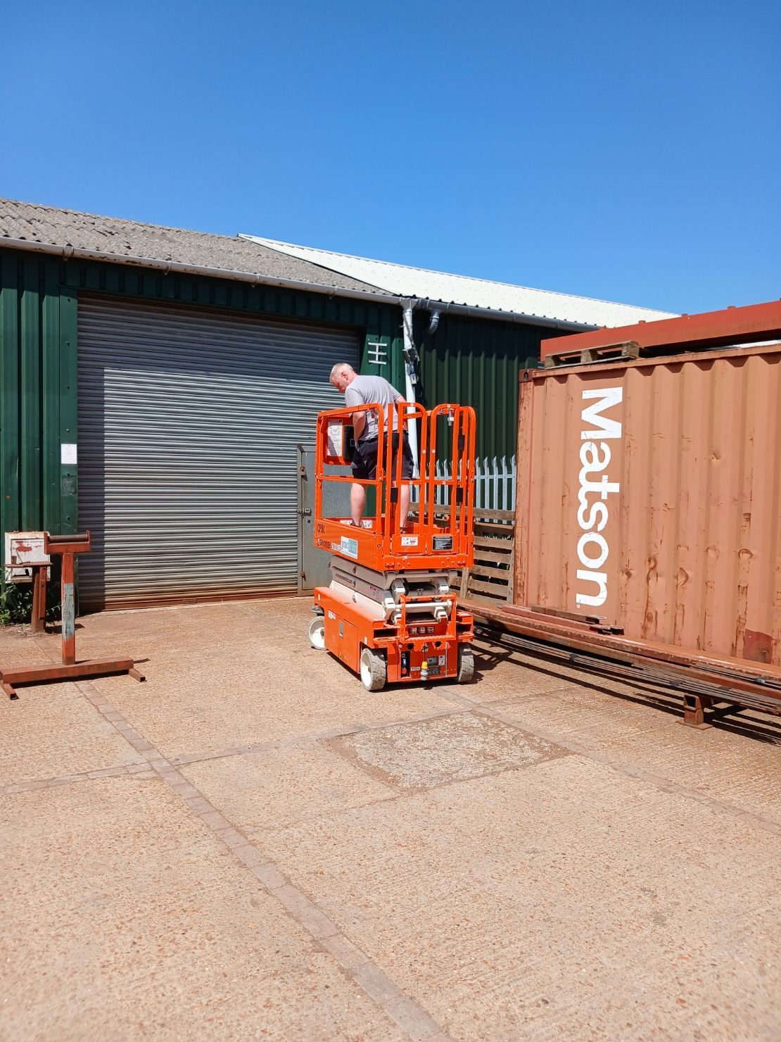 Orange scissor lift beside a shipping container outside a warehouse under a clear blue sky