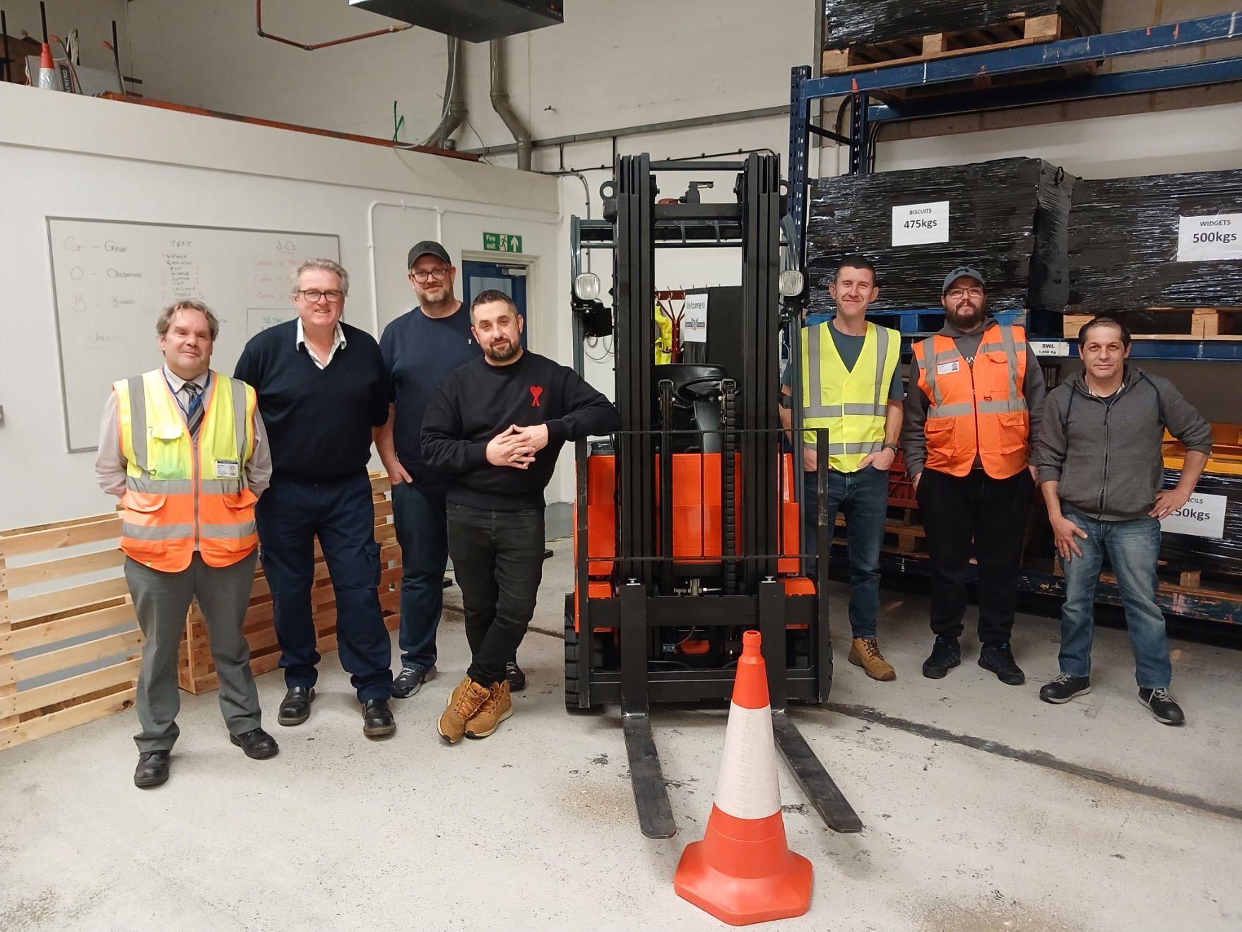 Six people posing in a warehouse beside a forklift and orange safety cone.