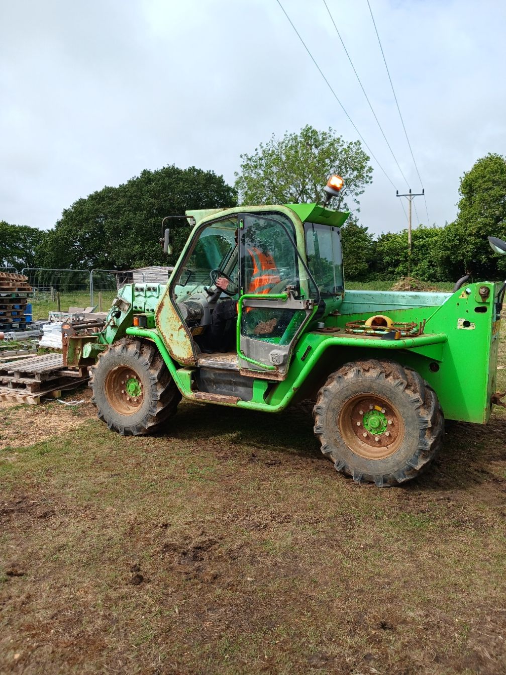 Green telehandler parked on muddy ground at a work site, with trees and equipment in the background