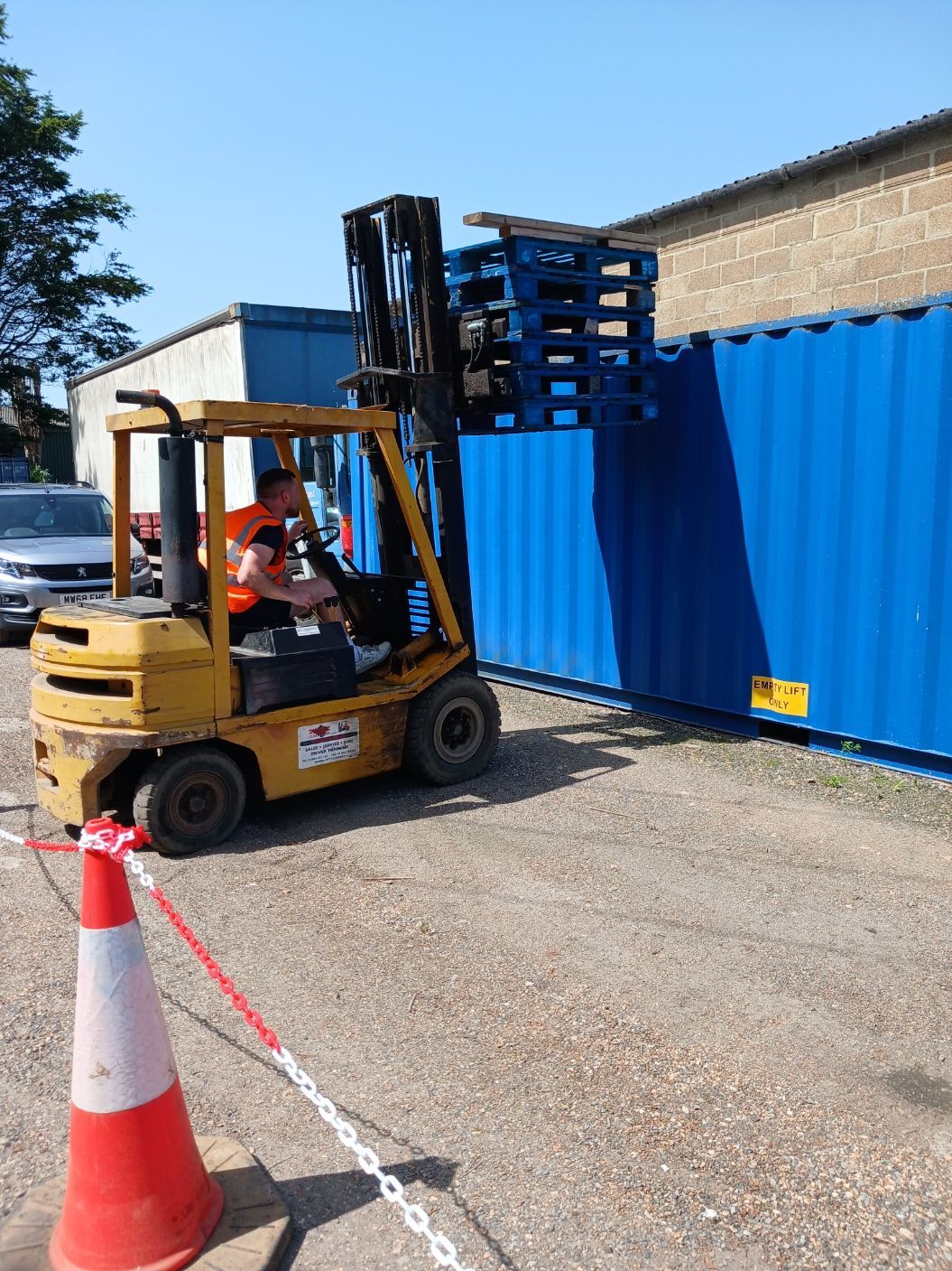 Forklift loading stacked pallets beside a blue shipping container outdoors