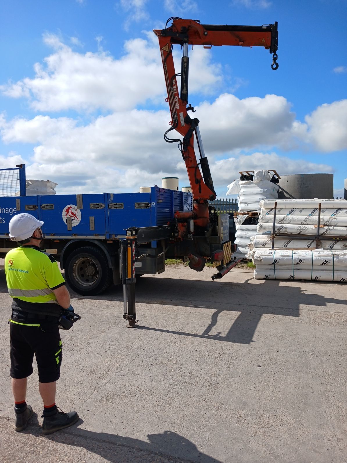 Worker watching a crane unload wrapped materials from a truck in an industrial yard