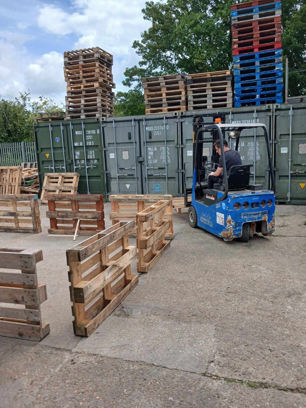 Forklift moving wooden pallets beside stacked shipping containers in an industrial yard.