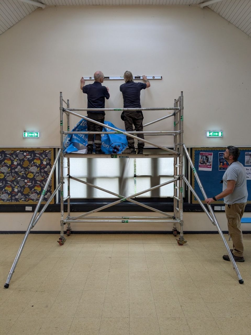 Two workers on scaffolding level something on a wall; another steadies the scaffolding. Inside a room.