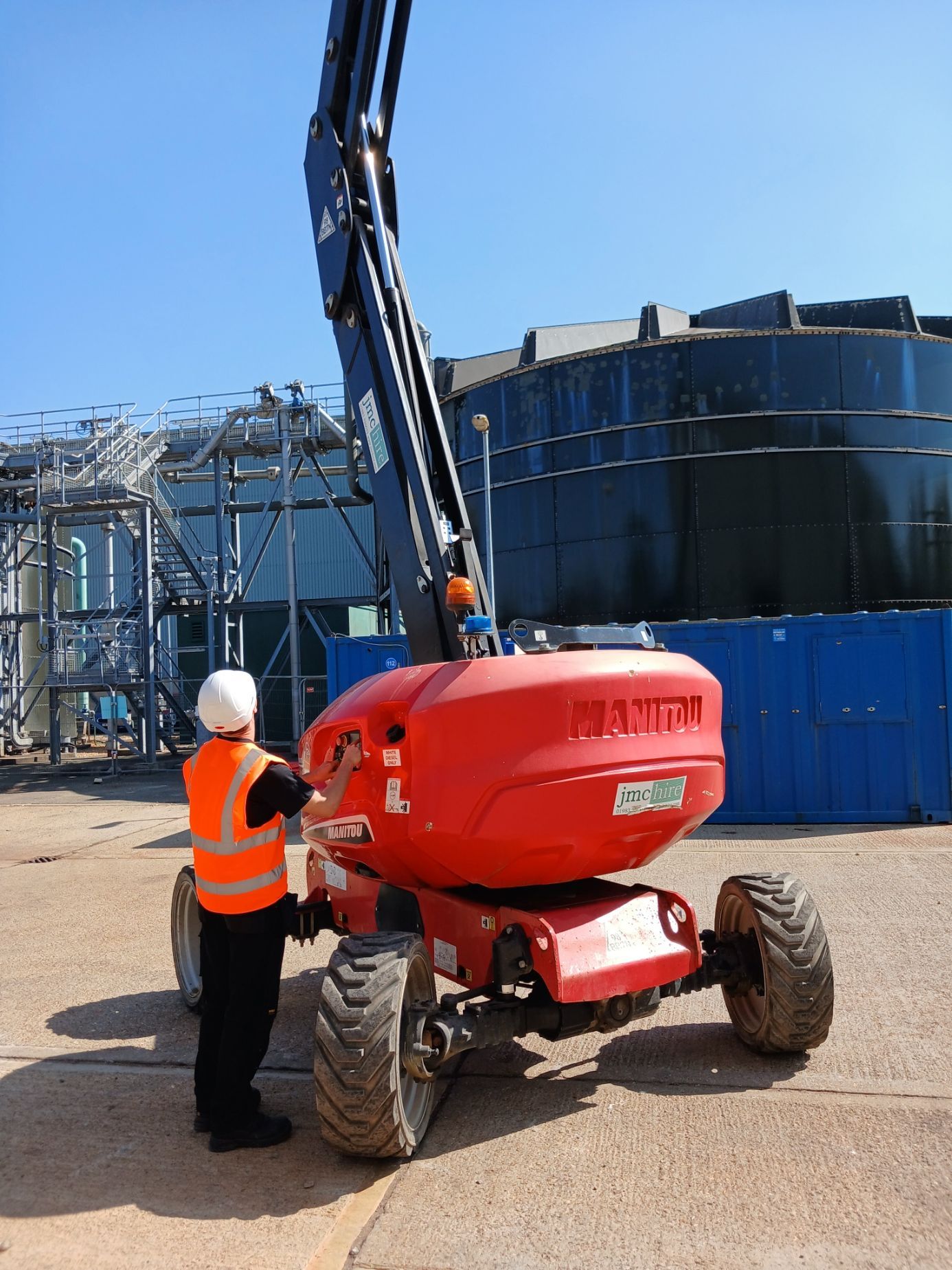 Worker in orange vest and hard hat beside a red boom lift at an industrial site.