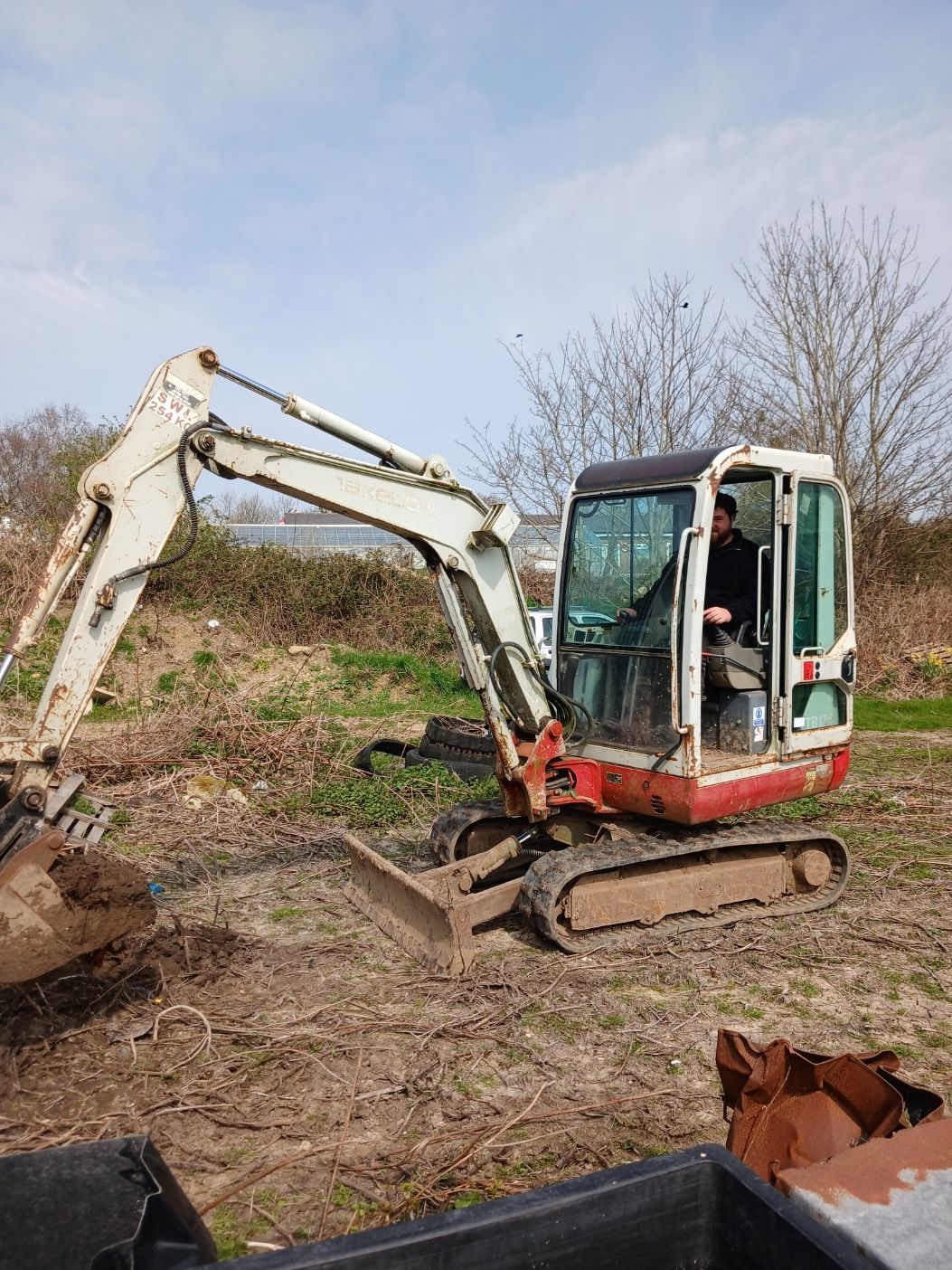 Small red excavator digging in a muddy field beside a brush pile under a cloudy sky