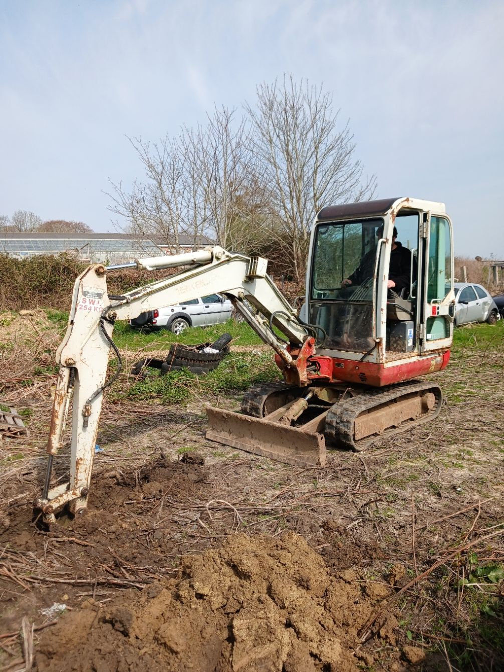 Excavator digging muddy ground beside a pond, with a white car in the background.