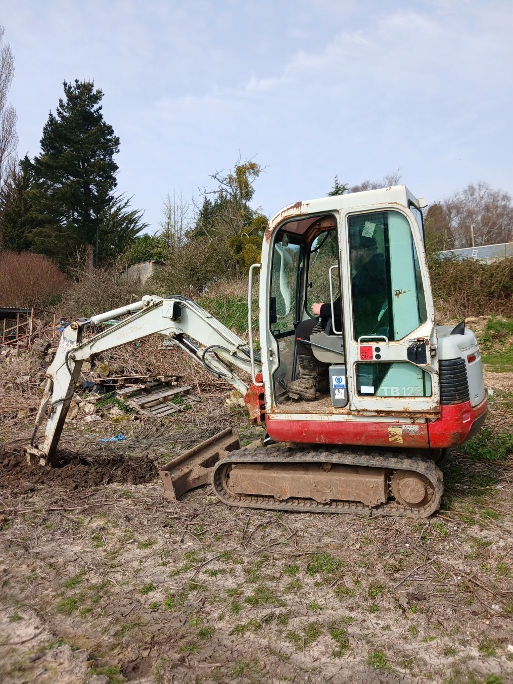 Small excavator on muddy ground beside a pile of brush and trees in a rural yard