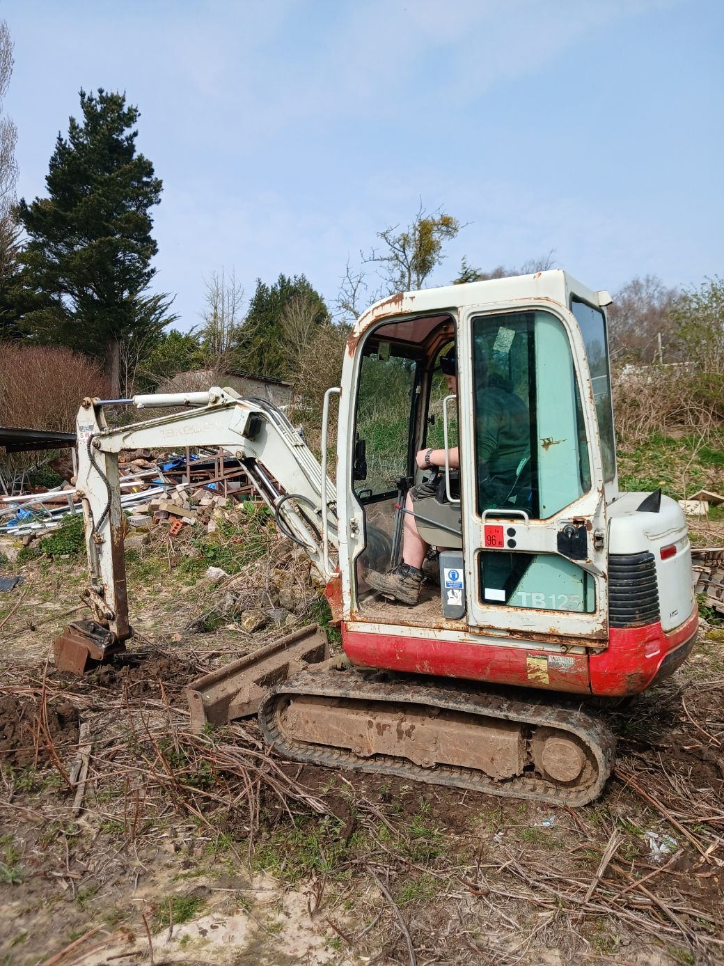White mini excavator on tracks digging in a muddy, brush-filled field under a blue sky.