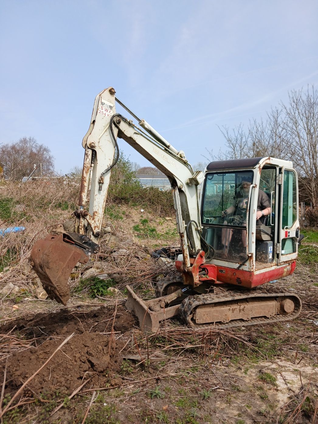 Excavator digging soil on a brush-covered hillside under a blue sky