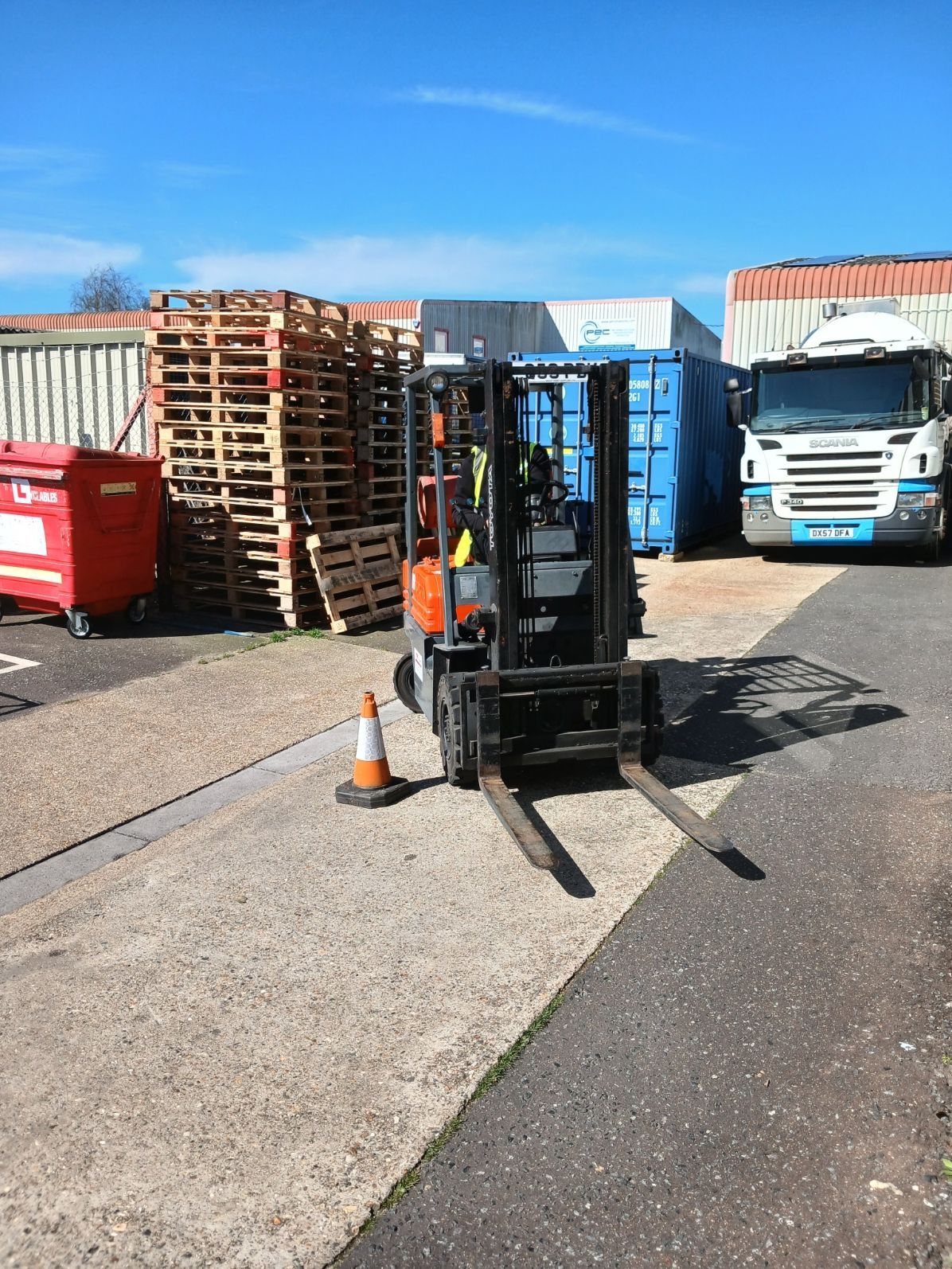 Forklift parked in an industrial yard beside stacked pallets, dumpsters, and parked trucks.