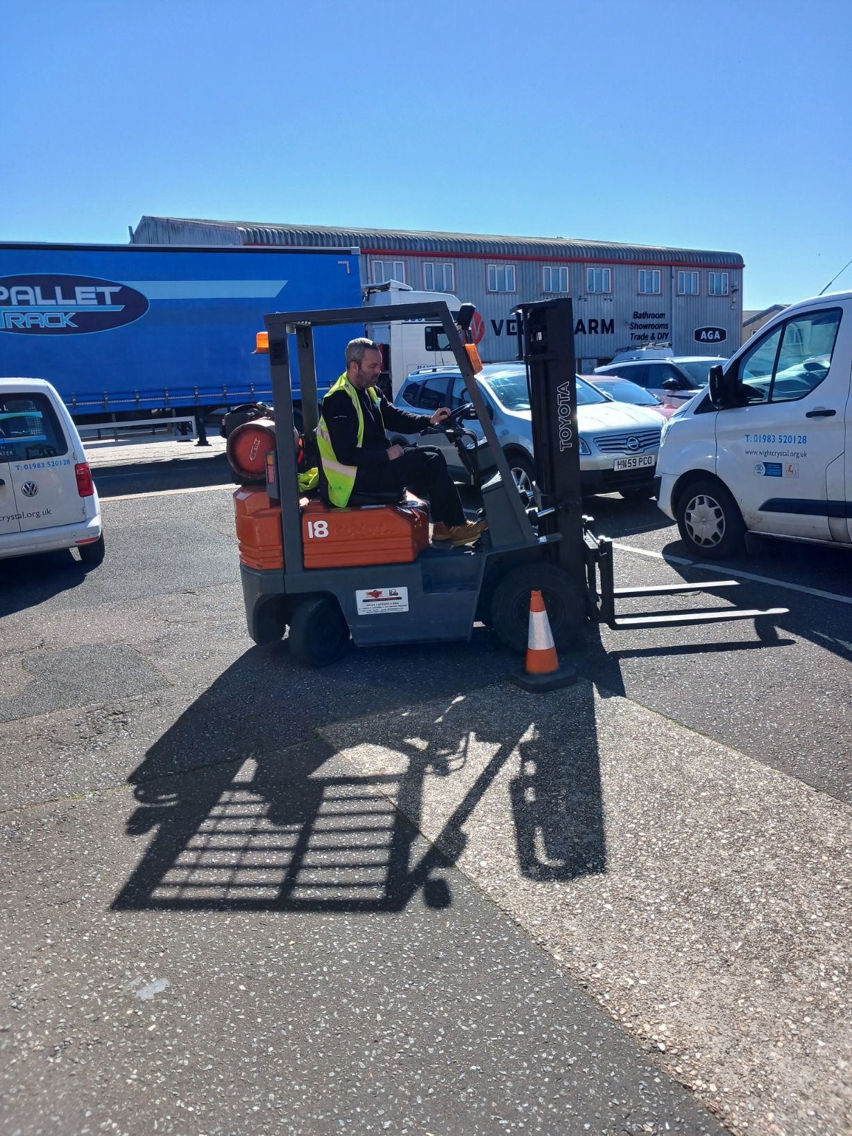 Forklift operator in safety vest moving a pallet outdoors beside parked vehicles and a blue trailer.