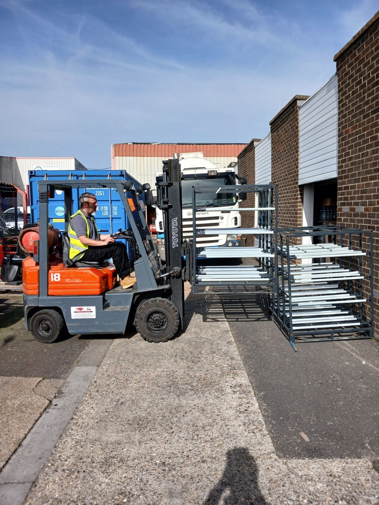 Forklift loading stacked metal racks in an industrial yard between brick buildings.