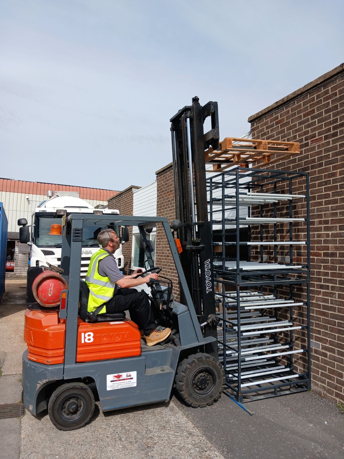 Forklift driver loading stacked metal racks beside a brick building