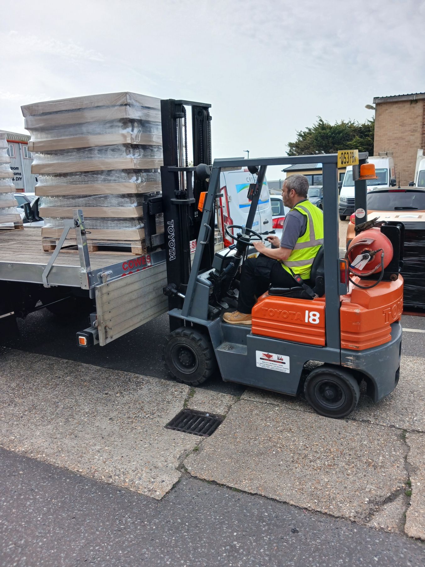Worker driving orange forklift carrying stacked pallets on a street beside a building