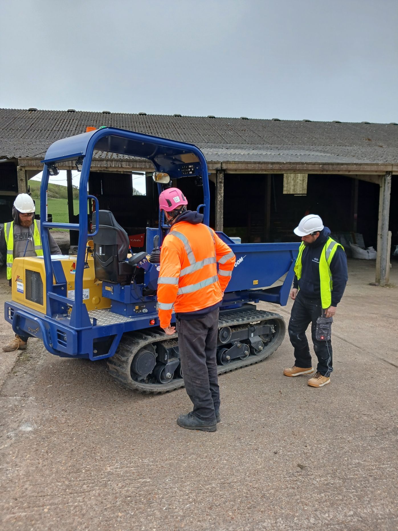 Construction workers inspect a blue mini excavator on a gravel site in front of a building.