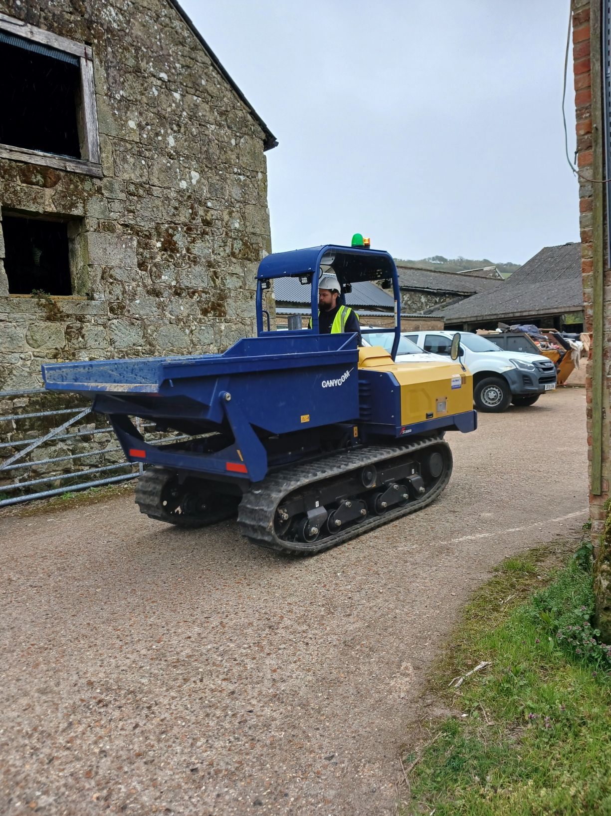 Yellow and blue tracked dumper on a gravel lane beside stone buildings, with a worker inside