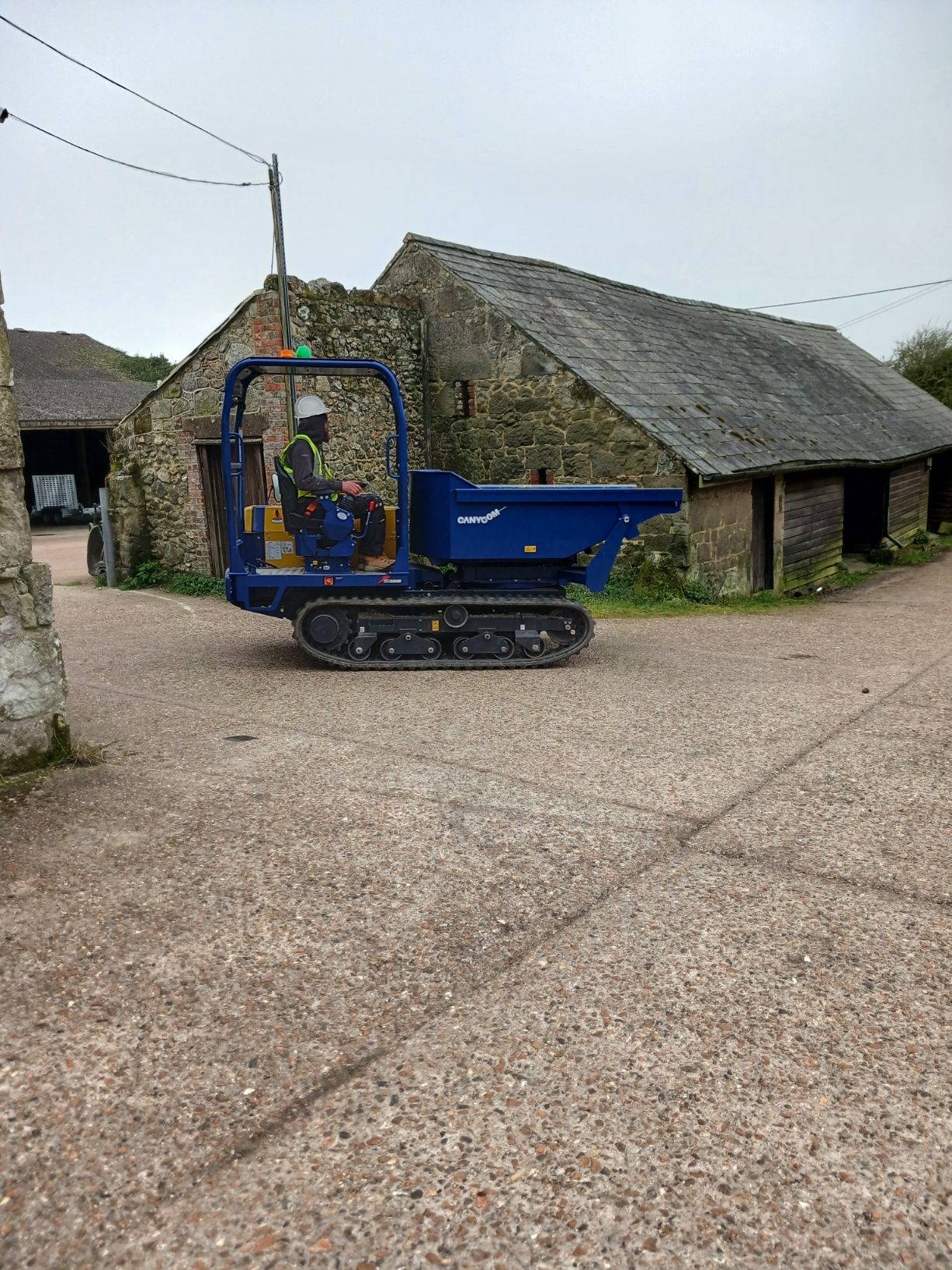 Blue mini dumper on a gravel farmyard beside stone barns under an overcast sky.