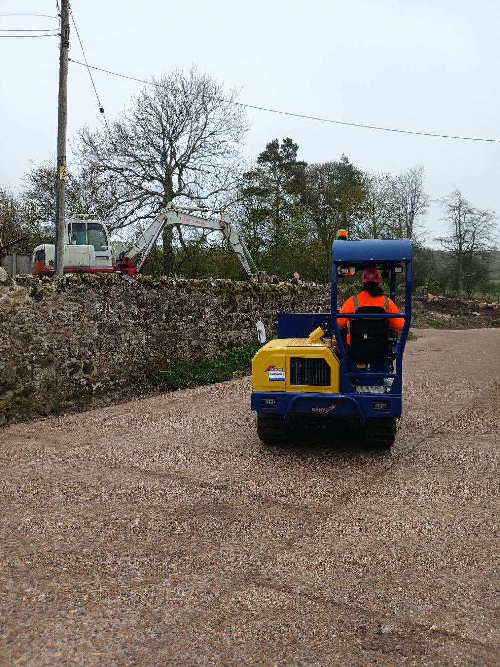 Blue utility vehicle with worker in orange vest on a gravel road beside a stone wall