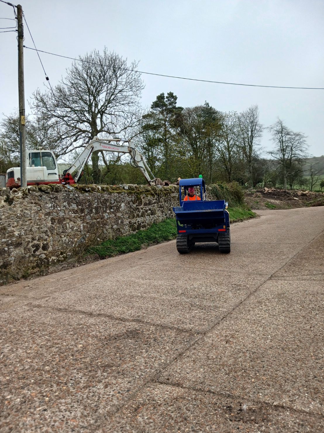 Small blue tractor driving on a gravel road beside a stone wall and trees under a gray sky