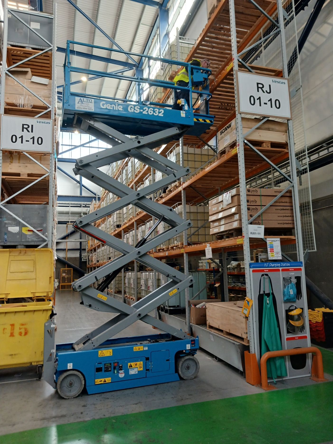 Blue scissor lift with worker raised in warehouse aisle between tall shelves and signs