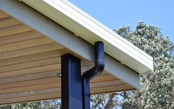 Black rain gutter and downspout on a beige house roof overhang against blue sky
