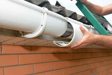 Hands install a white gutter on a brick house roofline.
